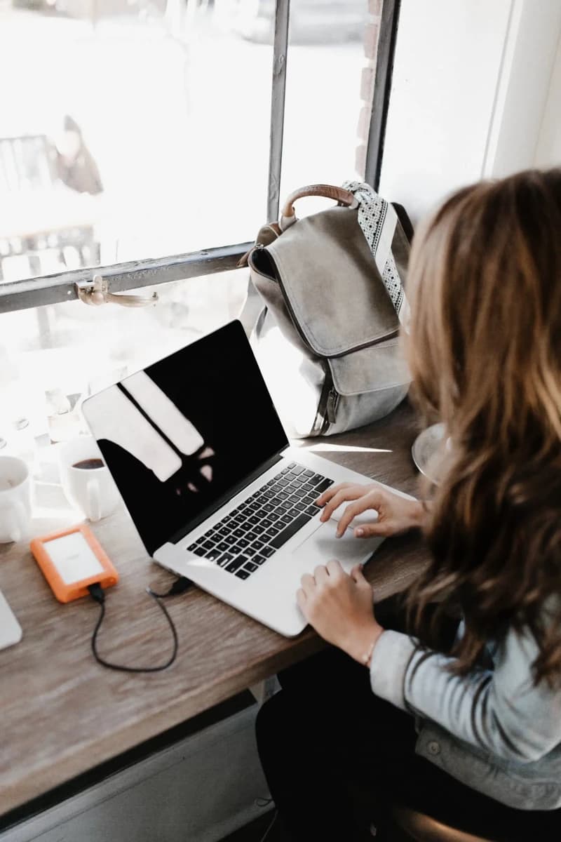 A woman with long brown hair is seated at a wooden table by a large window, typing on a silver laptop with a black screen displaying a white abstract graphic; beside her is a gray backpack with a patterned strap, an orange portable hard drive connected via cable, and a white coffee mug, all bathed in soft natural light suggesting a cozy, productive workspace in a café or home office environment.